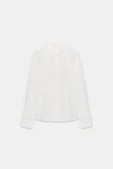 Camisa blanca de manga larga con cuello redondo tipo bebé y cierre frontal de botonadura simple.
