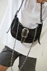 Cartera tipo baguette negra de cuero vacuno con textura tipo piel de cocodrilo. Tiene correa de cuero ajustable con detalles de tachas y cadena metálica plateada. El frente presenta un aplique de cuero con hebilla metálica plateada con tachas.