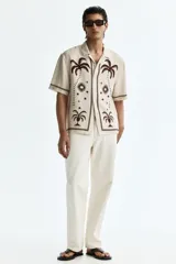 Camisa de manga corta color beige, con cuello abierto y cierre frontal de botones. Presenta un estampado en marrón de palmeras, soles y estrellas, con líneas en los bordes.