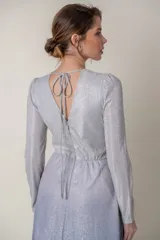 Vestido largo de lurex con acabado brillante en tono lila, con escote en V profundo, mangas largas y cintura marcada con un aplique bordado. La falda presenta un diseño drapeado que nace desde la cintura.