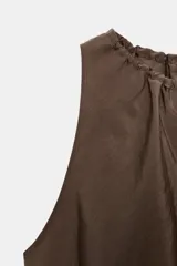 Vestido midi de color marrón con acabado satinado, cuello redondo con detalle fruncido y diseño sin mangas. Presenta un corte fluido con bajo asimétrico y bolsillos laterales ocultos.