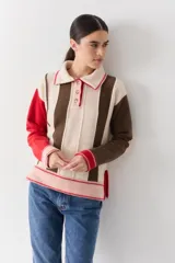 Buzo de punto con cuello polo y cierre frontal de tres botones. Presenta un diseño a rayas verticales en tonos beige y marrón, con mangas en bloques de color rojo y detalles en rosa en los puños y el ruedo.