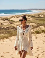 Vestido de playa corto en tejido calado color crema, con mangas largas acampanadas y diseño asimétrico con volado en el ruedo. Presenta un escote redondo con abertura frontal y cordón ajustable.