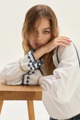 Blusa blanca de algodón con cuerpo de jersey y mangas de popelina. Presenta detalles de punto smock en puños y cuello, adornados con delicados bordados en azul y dorado.