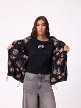 Campera reversible de diseño holgado, con un lado en tejido sherpa estampado con motivos florales en tonos negro y beige, y el otro lado en nylon negro. Cuenta con cierre frontal y cordones ajustables en el ruedo.