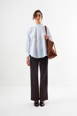 Blusa de algodón blanco con diseño de rayas verticales y textura de pequeños puntos en relieve. Presenta cuello alto con volados, cierre frontal con botones forrados y mangas largas abullonadas con puños ajustados.