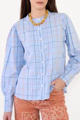 Camisa de manga larga con estampado de cuadros en tonos celestes, naranjas y verdes. Presenta un diseño con pliegues verticales en el frente, cuello redondo y cierre frontal con botones. Las mangas tienen un ligero frunce en los hombros y puños abotonados.