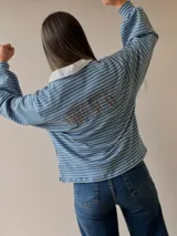 Buzo tipo chomba de corte oversize con diseño a rayas horizontales celestes y blancas. Presenta cuello camisero blanco y un bordado en la espalda con la inscripción República Oriental del Uruguay.