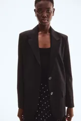 Blazer largo negro, de corte recto y botonadura sencilla con dos botones frontales. Presenta solapas de muesca, bolsillos delanteros ribeteados con solapa y hombreras.