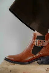 Botineta de cuero charol negro con acabado brillante, diseño de punta fina y taco cuadrado. Presenta un panel elástico lateral y una correa decorativa con hebilla metálica plateada.