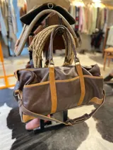 Bolso de viaje de cuero marrón oscuro con detalles en gamuza tostada, doble asa de mano y correa ajustable para el hombro.