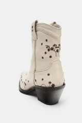 Botín estilo cowboy confeccionado en piel color crudo, con diseño de flores bordadas en tono marrón. Presenta tacón ancho en bloque, acabado en punta y tiradores laterales.