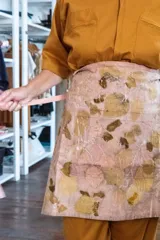 Delantal corto de algodón color beige con estampado de hojas en tonos marrones y amarillos. Tiene un bolsillo frontal y se ata a la cintura con una cinta fina color rosa.