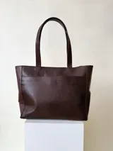 Cartera tipo tote de cuero graneado negro, con diseño clásico y textura uniforme. Cuenta con doble asa de hombro, bolsillo frontal con cierre magnético, bolsillo trasero con cierre metálico y compartimento principal con cierre. El interior está forrado y dispone de múltiples bolsillos organizadores.