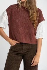 Chaleco sin mangas de tejido punto, color terracota o rojizo oscuro, con textura visible y brillo sutil (lurex). Se lleva superpuesto sobre una camisa blanca de manga corta con detalles de encaje o bordado en las mangas.