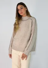 Sweater de punto en color beige melange, con cuello alto y costuras decorativas visibles en contraste a lo largo de las mangas y hombros. Presenta un diseño de corte holgado con puños y ruedo acanalados.