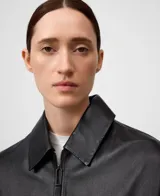 Chaqueta de piel ovina color negro con diseño desestructurado y holgado. Cuenta con cuello de solapa, cierre frontal de cremallera, bolsillos de vivo inclinados en el delantero y una capelina en la espalda que aporta volumen.