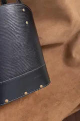 Cartera matera de cuero vacuno negro con textura símil lagarto, diseño estructurado con base ovalada y doble asa de hombro. Cuenta con detalles de tachas metálicas doradas y costuras pespunteadas en contraste.