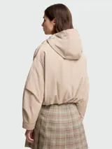 Campera corta con capucha, color beige, de corte holgado y diseño minimalista. Presenta cierre frontal oculto, bolsillos laterales con solapa y botones a presión, mangas largas con puños ajustables y cordones decorativos en la capucha. Incluye el logo de la marca en la manga izquierda.