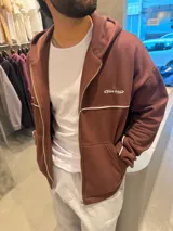 Campera con capucha de corte boxy color marrón, con cierre frontal completo y bolsillos tipo canguro. Presenta una línea blanca horizontal en el pecho que se extiende por las mangas y un pequeño bordado con texto en el pecho.