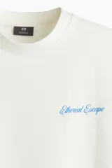 Remera color hueso de corte oversized, confeccionada en tejido de algodón pesado. Presenta cuello redondo acanalado, hombros caídos y estampado pequeño con la frase "Ethereal Escape" en el lado izquierdo del pecho.