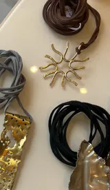 Collar artesanal con dije de diseño orgánico en metal y tiento de gamuza ajustable. El dije presenta una forma circular con terminaciones curvas, evocando un diseño natural.