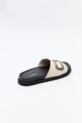 Sandalia slide color beige con hebilla metálica plateada y suela negra.