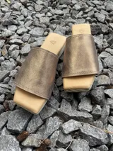 Zueco de mujer con plataforma de madera y suela de goma. La capellada es una tira ancha de cuero dorado metalizado, y la plantilla es acolchada.