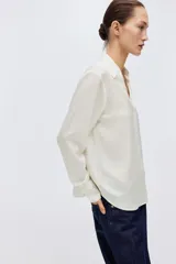 Blusa blanca de tela con brillo, cuello clásico, escote en V con botones frontales, mangas largas con puños sencillos y bajo redondeado.