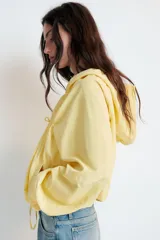 Chaqueta de nylon color amarillo pastel, con cuello subido y capucha. Presenta cierre frontal con cremallera, mangas largas con puños elásticos y detalle de lazadas ajustables en el frente. El bajo cuenta con cordón elástico y stoppers para un ajuste personalizado.