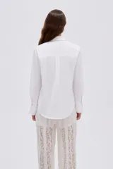 Camisa blanca de algodón, holgada, de manga larga con puños anchos, cuello camisero y cierre frontal oculto con un botón dorado visible en el cuello.