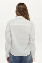 Camisa blanca de manga larga abullonada con detalle de punto smock en los puños y cordones para atar. Presenta cuello clásico y cierre frontal de botones.