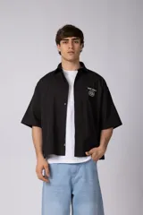 Camisa de manga corta negra, de corte boxy, con cuello camisero, cierre frontal de botones y bordado circular en el pecho.