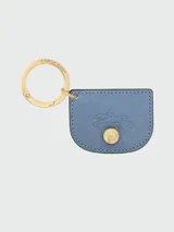Llavero Longchamp de la línea Épure, confeccionado en cuero azul con textura granulada. Presenta una forma semicircular con el logo de la marca grabado y un botón metálico dorado con el emblema de Longchamp. Incluye una argolla metálica dorada grabada con el nombre de la marca.