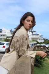 Chaqueta de gamuza color beige con diseño de flecos en el frente y las mangas. Presenta solapas clásicas, cierre frontal con botones y bolsillos laterales.