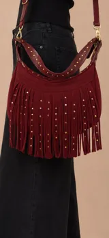 Cartera bandolera color bordó de gamuza con flecos largos y apliques de tachas doradas. Incluye una correa corta decorada con tachas y una correa larga extensible y extraíble, ambas con herrajes metálicos dorados.