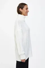 Polerón largo de tejido de punto en color blanco, con cuello alto y diseño de trenzas verticales. Presenta un corte amplio y mangas largas con puños acanalados.