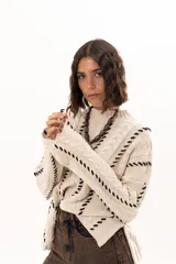 Buzo de tejido de punto color crudo con diseño de trenzas y costuras decorativas en color negro que recorren la prenda en forma de espiral. Presenta cuello redondo y mangas largas con terminación acampanada.