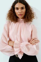 Mono largo con parte superior de blusa rosa de manga larga, cuello alto fruncido y puños con volantes. La parte inferior es un pantalón negro de pernera ancha con bolsillos delanteros y un cinturón negro con hebilla dorada.