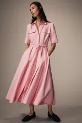 Vestido midi de estilo camisero color rosado, con cuello solapa, manga corta y cierre frontal con botones. Presenta bolsillos de plastrón con solapa en el pecho, bolsillos laterales ocultos y un cinturón del mismo tejido para ajustar en la cintura.