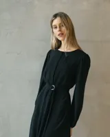 Vestido midi negro de silueta fluida, con mangas largas abullonadas y cintura marcada mediante un cinturón incorporado con hebilla metálica. Presenta un diseño de escote cerrado y caída liviana.