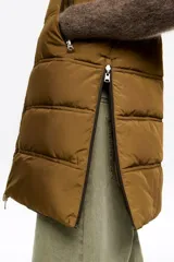 Chaleco acolchado de largo medio y corte en línea A, confeccionado en tejido técnico color beige con detalles en negro. Presenta cuello subido, capucha desmontable, cierre frontal de cremallera, bolsillos laterales con cremallera, logo bordado en el pecho y aberturas laterales con cremallera.