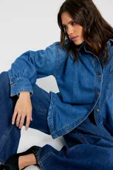 Camisa de denim azul de calce amplio, con cuello clásico, botonadura frontal, bolsillo plaqué en el pecho, hombros caídos y puños anchos abotonados.