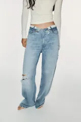 Jeans de mezclilla de tiro medio, corte wide leg y diseño relaxed. Presenta detalles rotos y desgastados en las piernas, y una cinturilla doblada superpuesta con trabillas y cierre frontal con cremallera y botón metálico.