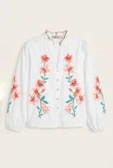 Camisa blanca de manga larga abullonada, con delicados bordados florales en tonos azules y verdes a lo largo de la botonadura frontal y en las mangas. Presenta cuello mao con reborde en contraste y cierre frontal con botones.
