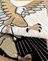 Remera blanca de algodón con cuello redondo y manga corta. Presenta un bordado de un águila con lentejuelas doradas, plateadas y negras en el frente.