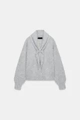 Jersey de punto gris con cuello tipo pañuelo integrado, manga larga con puños ajustados y aplicación metálica en el cierre frontal.
