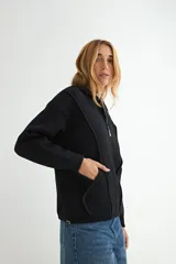 Abrigo negro con capucha, confeccionado en tejido de textura suave. Presenta cierre frontal con cremallera, bolsillos laterales integrados con costuras decorativas y puños ajustados.