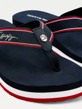 Ojotas de mujer marca Tommy Hilfiger con suela de cuña. Cuentan con tiras de nailon trenzado en color azul marino con detalles en rojo y blanco, firma de la marca bordada y logo metálico. La suela de goma presenta franjas decorativas en los colores distintivos de la marca.