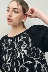 Vestido corto negro con bordado floral blanco, escote redondo con abotonadura frontal y mangas 3/4 abullonadas con pliegues en los hombros.